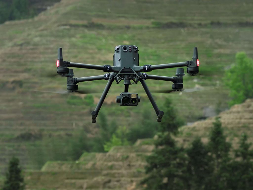 Drone DJI Matrice en vol avec caméra et lumières rouges, sur fond de collines vertes.